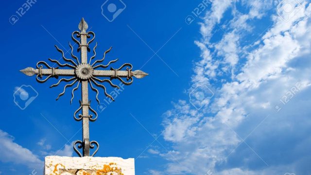 Stone roof of church with wrought iron cross against the blue sky